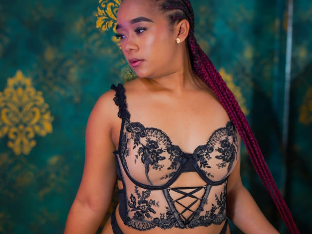 KailaniMoani with maroon braids, wearing a black lace bra that shows her tits, has a cute face, gold earrings, and a sexy look. Her tits are medium-sized.