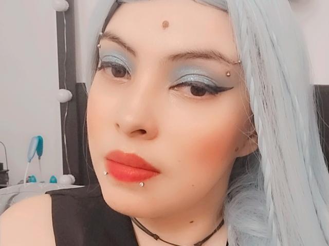 NatashaYou69 with pale skin, blue eyeshadow, red lips, silver hair, facial piercings, lip piercing, and a black choker. Her tits are small, and she's looking at the camera.