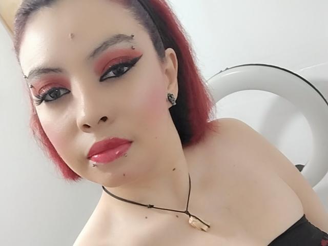 NatashaYou69 has red eyeliner, red lipstick, and red eyeshadow with small piercings on her nose and lip. She has red hair and a black strapless top.