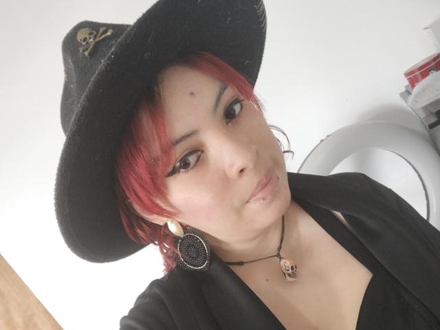 NatashaYou69 with red hair, black hat, skull button, black jacket, and circular earrings, pouting slightly, wearing a skull necklace, looking at camera.