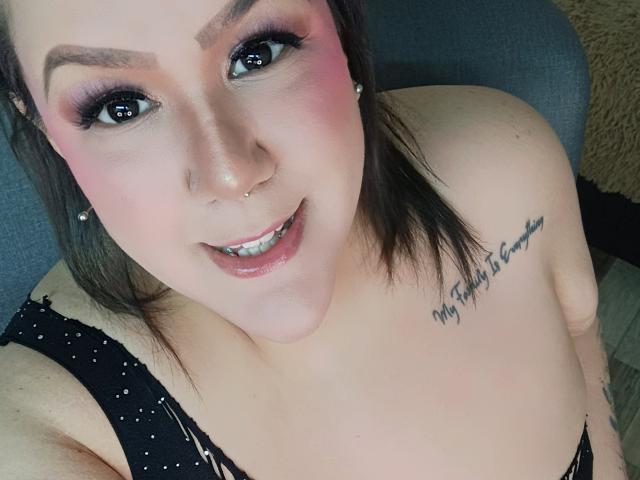 GiiannaMorrys has pink eyeshadow, black eyeliner, nose ring, smiling with white teeth, black polka-dot top, tattoo on right shoulder, and brown hair, looking up at the camera.