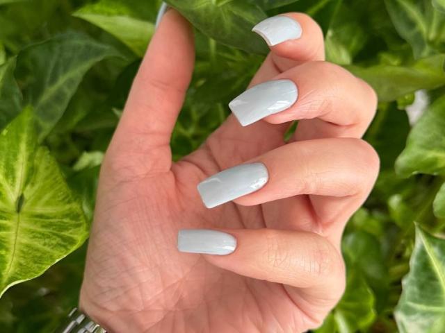 Teffi's hand with shiny, pale gray, almond-shaped nails, showing off her manicure against green leaves. Her skin is light, and she's wearing a silver bracelet.