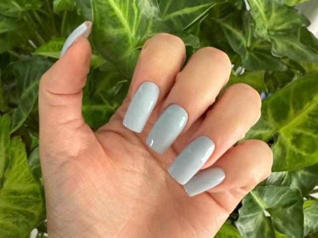 Teffi's hand with pale, glossy, long, square-shaped nails. She's got a light skin tone and the nails are a shiny, pastel blue. Green leaves are in the background.