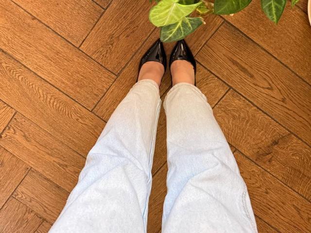 Teffi's legs in light blue jeans and black pointed shoes, standing on wooden floor, with green leafy plant in the top right.
