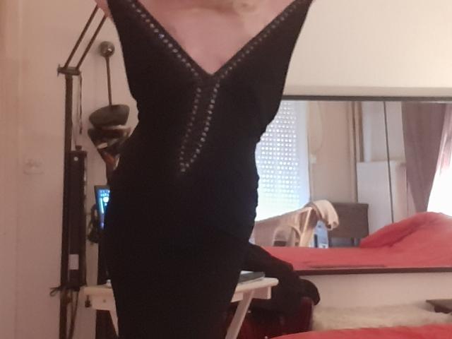 IssahneMay in a tight black dress with a deep V-neck and silver studs, standing with arms raised, showing her ass and small tits. Bedroom setting.
