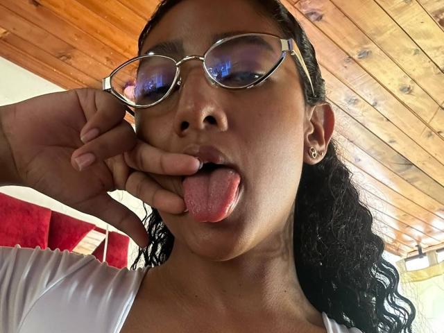 NatalyBlonder with dark curly hair, wearing glasses, sticking her tongue out, and touching her lips with her finger. She has a small nose and is wearing a white top.