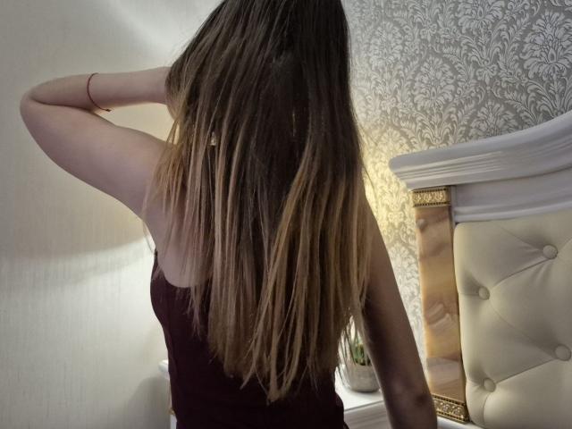 Seliina's ass-up, long brown ombré hair, wearing a black tank top, hand in hair, standing in front of a white, tufted bed.