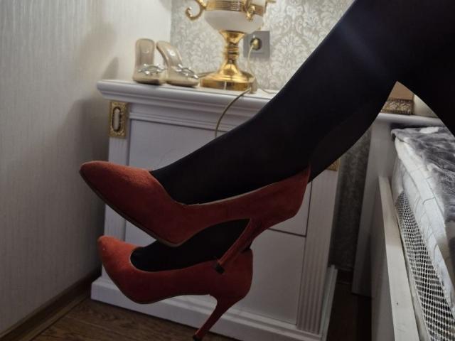 Seliina's black legs in red suede high heels with black stockings, crossed at the ankle, resting on a white wooden dresser with a gold lamp and beige shoes.