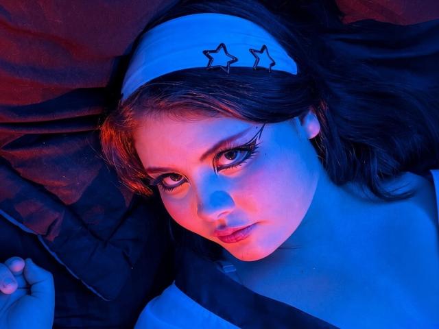 MicheMeow has blue and red neon lighting on her face, with star-shaped headband, black eyeliner, and looking up with serious expression, lying on dark fabric.