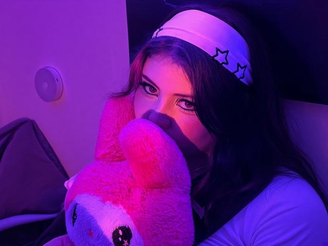 MicheMeow with dark hair, white headband, and pink mask, holding pink teddy bear, purple and pink lighting, looking at camera, wearing white top, serious expression.