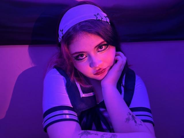 MicheMeow with purple-blue lighting, head resting on hand, wearing a white headband with black star pattern, and a sailor-style shirt, has tattoos on her right forearm.