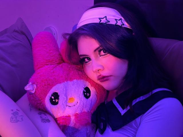 MicheMeow with purple-lit face, dark hair, and white headband, hugging pink fluffy toy, wearing white sailor shirt, resting on pillow, showing tattoo on left wrist.