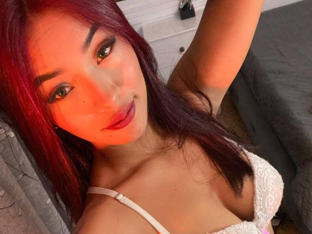 SashaHans with red hair, medium tits in white lace bra, orange skin, dark eyes, full red lips, arm raised, taking selfie, in bedroom, gray bed in background.