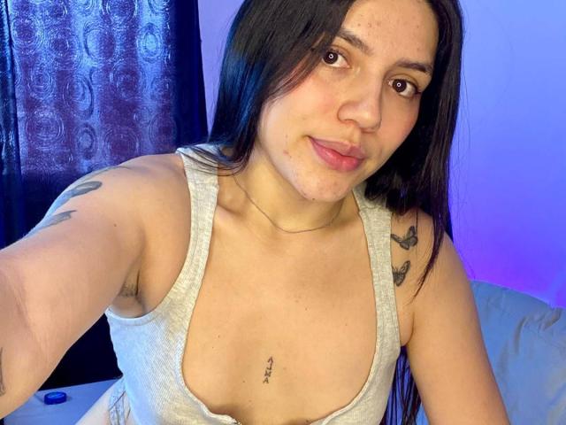 SophiaMarx with long black hair, light skin, small tits, gray tank top, butterfly tattoos, and a nipple piercing, smiling at the camera.