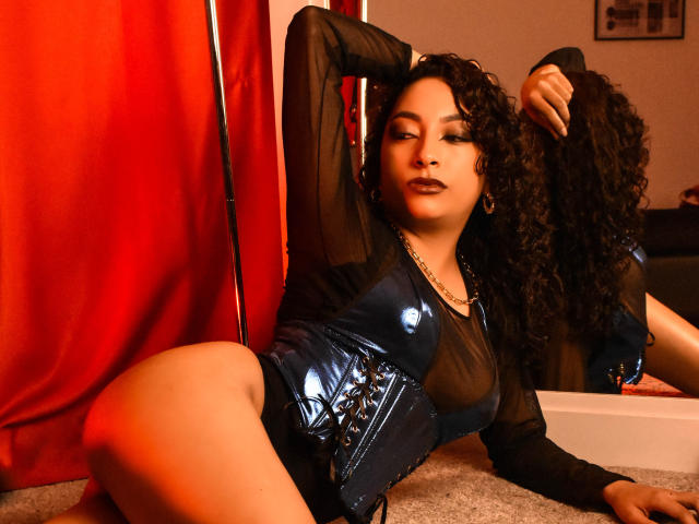 CrystalWright with curly black hair, wearing a sheer black top and shiny blue vinyl corset, lying on carpet, one arm behind head, reflecting in mirror, red curtain in background.