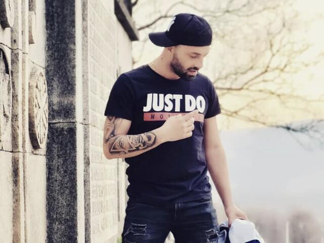 TommyDum with a black cap, black t-shirt, and black jeans, holding a white bag, has a tattoo on his right arm, looking down, standing by a stone wall.