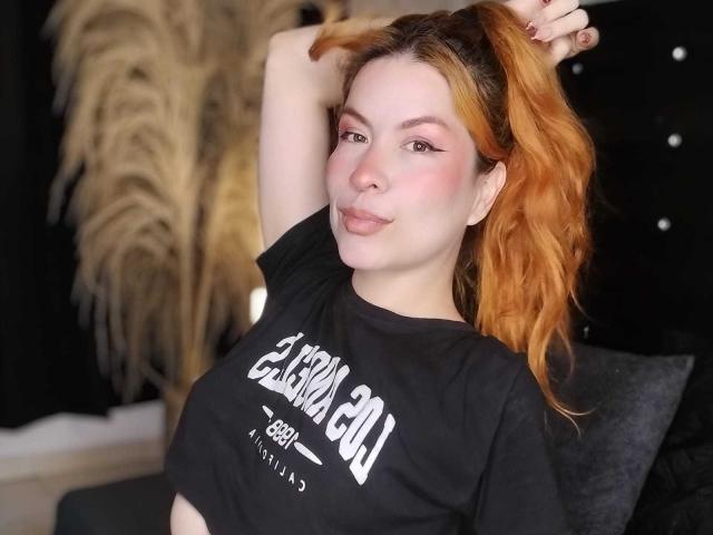 LilithSnop with orange ponytail, black t-shirt, pink eyeliner, and slight smile, holding ponytail, showing tits, fair skin, black background, and puffy pink cheeks.