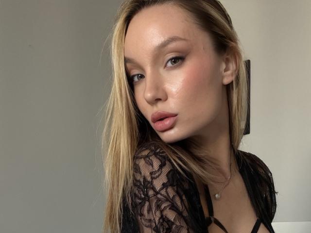 TitsQueen avec une peau éclatante, des lèvres roses et pulpeuses, et des cheveux blonds, portant un haut en dentelle noire, regardant séductrice l'appareil photo. Son maquillage est subtil, mettant en valeur ses traits attrayants..