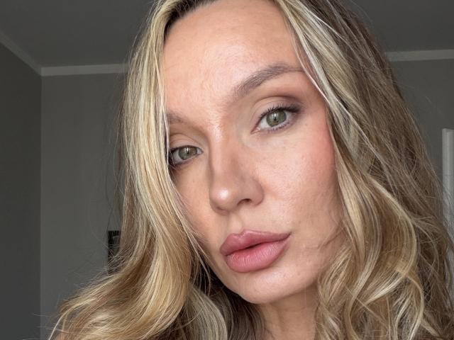 TitsQueen ha i capelli biondi e ondulati, gli occhi verdi e le labbra rosa. Il suo viso è leggermente cosparso di lentiggini e guarda direttamente la telecamera con un'espressione neutra..
