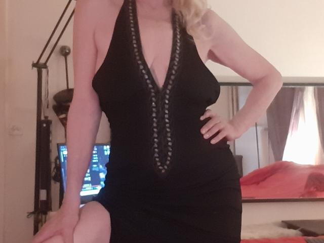IssahneMay trägt ein schwarzes Kleid mit tiefem V-Ausschnitt und verzierten Details, das ihre großen Brüste und ihren wohlgeformten Po zur Geltung bringt. Sie steht mit einem angewinkelten Knie da, hat blondes Haar und legt eine Hand an die Hüfte..