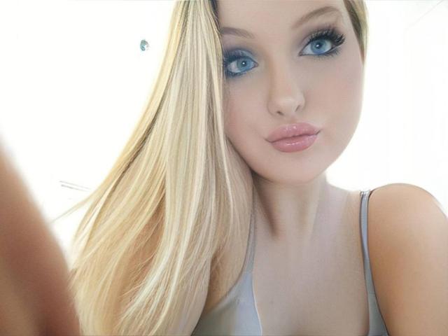 SimonaKorzep has blonde hair, blue eyes, and shiny pink lips. She's wearing a gray top, looking at the camera with a slight pout, and has long, straight hair.