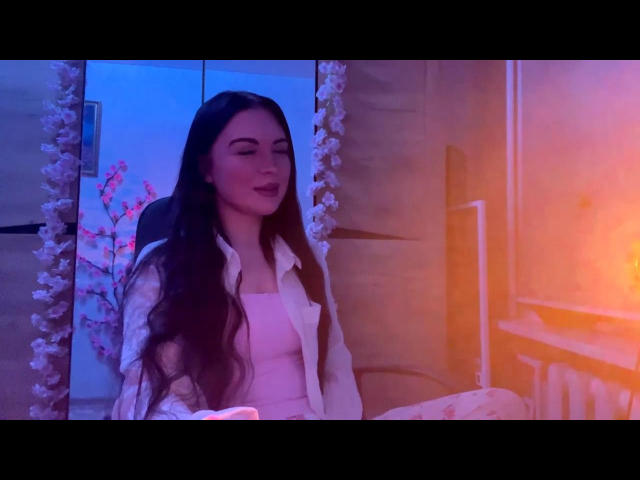 JerleneLeoni with long dark hair, white shirt, and pink top, sitting in a chair, eyes closed, pink and blue lighting, white floral curtain on the left.