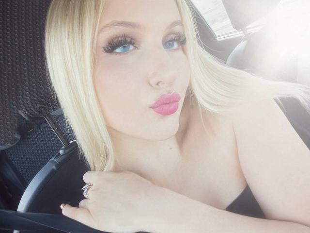 SimonaKorzep with blonde hair, blue eyes, and pink lipstick, pouting her plump, shiny lips, holding a black strap, wearing a ring, in a car.