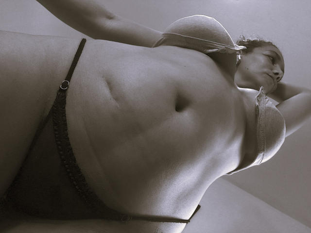 AmberDemons with big boobs, wearing a bra and black panties, arms raised, standing from a low angle, showing a belly button, and a slightly chubby, sexy body.