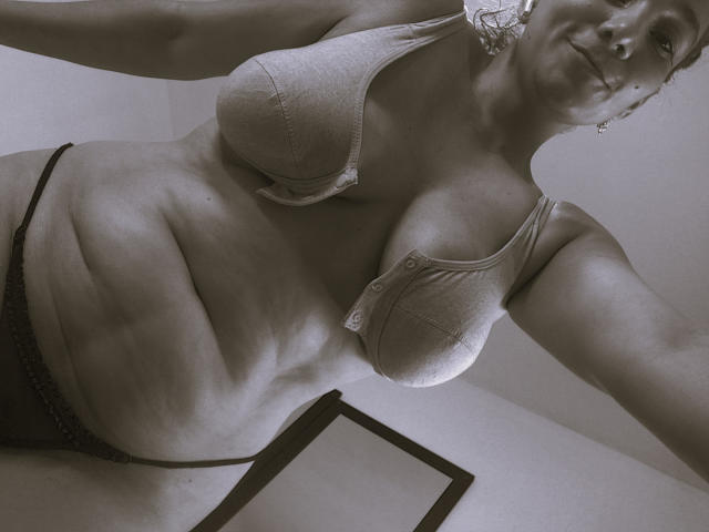 AmberDemons with large, saggy tits, wearing a buttoned bra and black panties, showing a chubby, muscular, and slightly sweaty belly, taking a selfie from a low angle.