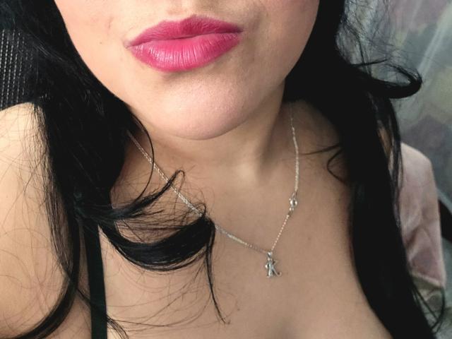 VivianaThompson's face with shiny pink lipstick, black wavy hair, and a silver necklace with a small key pendant, showing her cleavage. Her skin is light, and she's smiling.