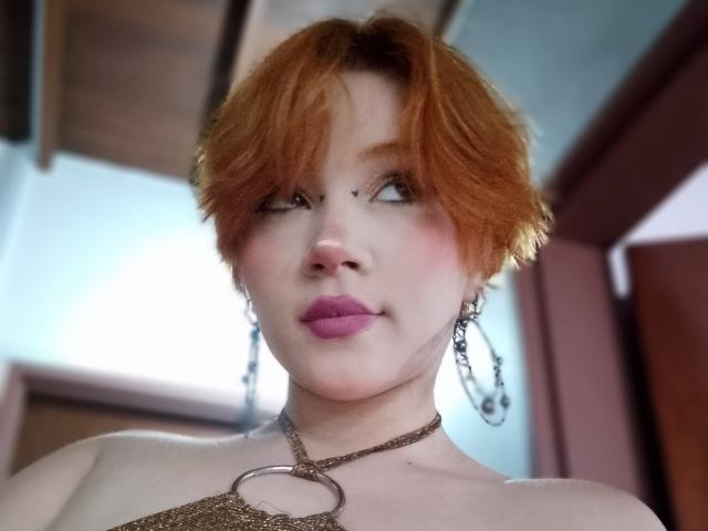 ChloeeWilliams has short red hair, pink lipstick, and a nose piercing. She's wearing a chain necklace and dangling earrings, with a subtle smile and soft makeup.