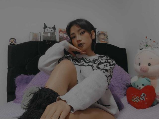 HenutmireRossi, con cabello negro corto, vestida con un suéter blanco y negro, sentada en una cama con almohadas moradas, botas negras y un cerdito de peluche con un corazón..