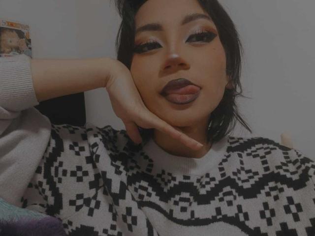 HenutmireRossi with dark makeup, tongue out, black patterned sweater, hand on cheek, looking at camera, wearing long eyelashes, short black hair, light skin, and a sexy expression.