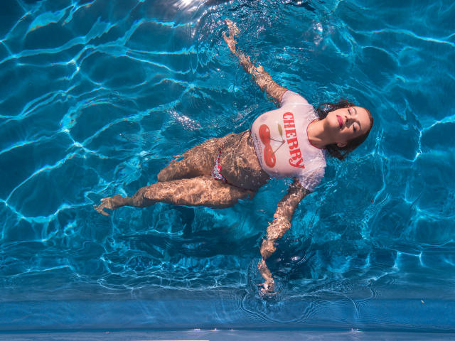 ScarletRouge è immersa in una piscina, indossa un top corto bianco di Hooters con il logo di una ciliegia rossa e dei pantaloncini da bagno rosa, con le braccia e le gambe distese e gli occhi chiusi..
