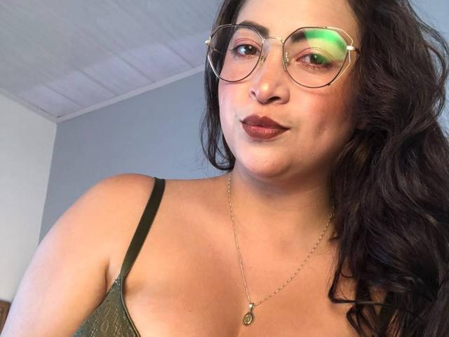 AlanaWest with glasses, dark wavy hair, green reflection in glasses, wearing a green spaghetti-strap top, necklace with a small pendant, brown lipstick, and a slight smile.