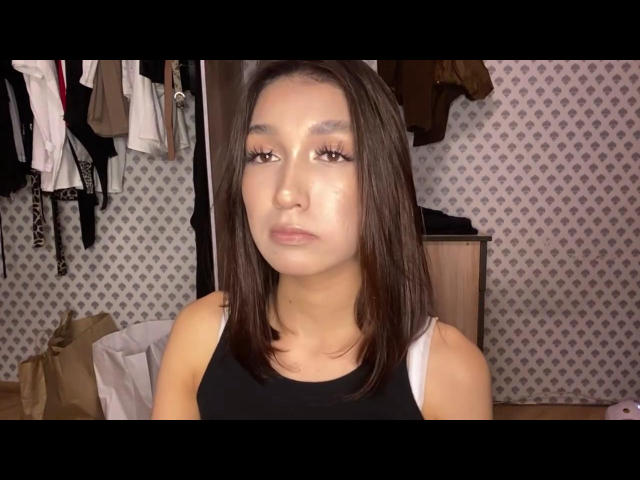 KalaMaynor has shiny brown eyes, medium-length dark brown hair, and a serious expression. She's wearing a black tank top, showing her small tits. Her ass is not visible.