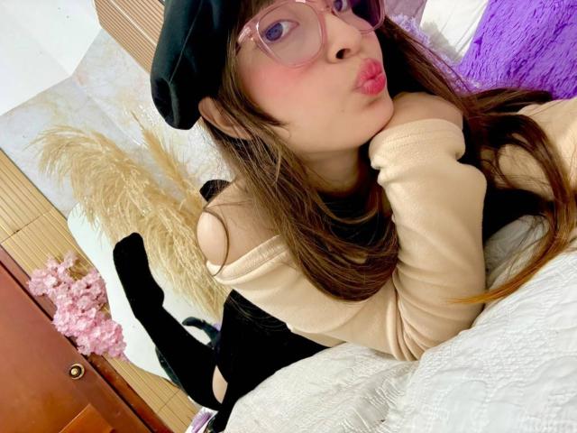 IsabelaP with pink glasses, black beret, pouty lips, beige top, black skirt, black socks, lying on white bed, brown hair, puffy cheeks, cute, soft lighting, bedroom.