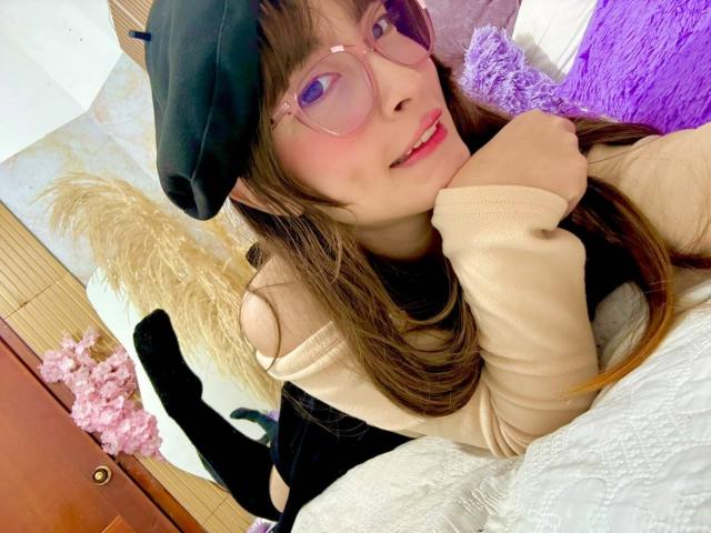 IsabelaP with glasses, black beret, and beige top, lying on bed, smiling, showing pink lipstick, black socks, and brown hair, hands under chin, looking at camera.