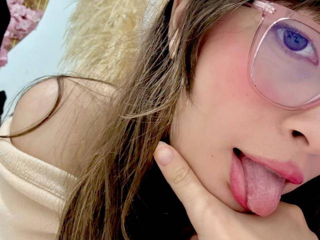 IsabelaP with pink-tinted glasses, tongue out, pink lips, and light brown hair, holding her cheek with her finger, showing a cute and playful expression.