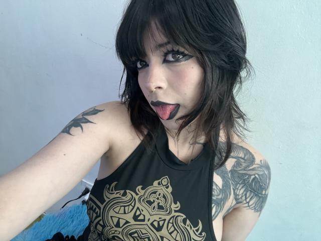 BabeSix has dark hair, black eyeliner, and black lipstick. She's sticking out her tongue, wearing a black top with gold patterns. She has tattoos on her arms.