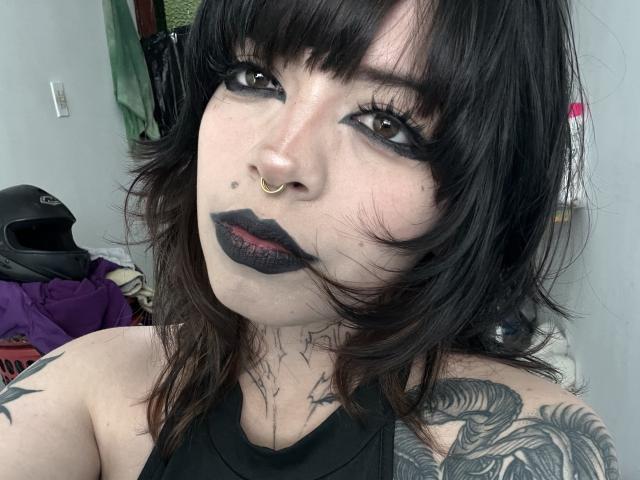 BabeSix with black lipstick, black eyeliner, and nose ring, has dark wavy hair, tattoos on shoulders, and a septum piercing, looking at camera with sultry expression.