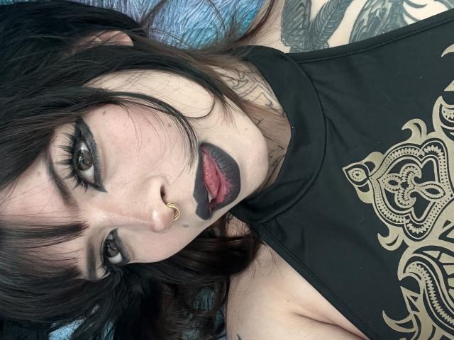 BabeSix with black lipstick, nose ring, and tattoos, lying down, wearing a black top with gold patterns, looking up with half-open red lips, and dark, wavy hair.