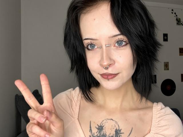 LianeBadenoch with black shaggy hair, blue eyes, nose ring, lip piercing, and a chest tattoo of a moon and stars, making a peace sign with her right hand.