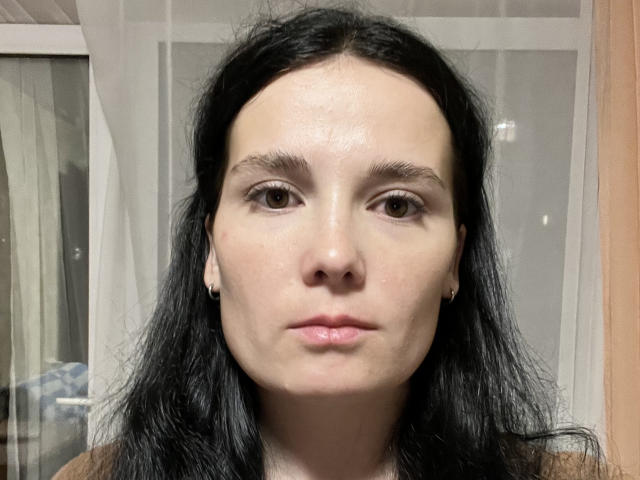 Heidy Heimbuch tem pele clara, cabelo preto ondulado, olhos castanhos e uma expressão neutra. Seus pequenos seios são visíveis sob uma blusa marrom. Ela está usando pequenos brincos..