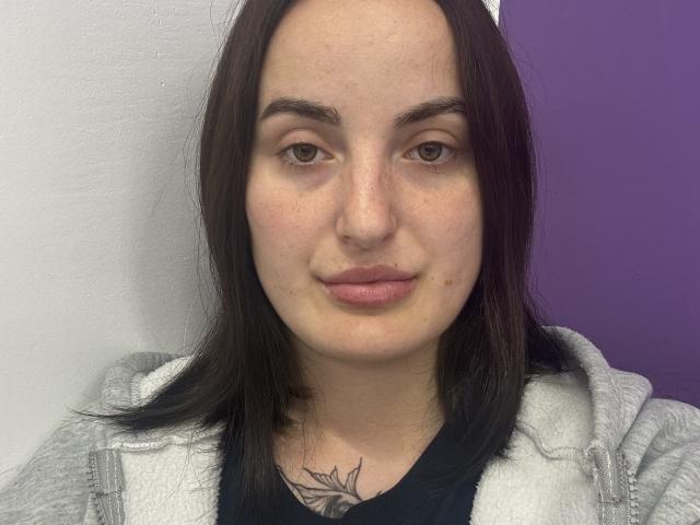 A GerriEdler tem um rosto adorável, com olhos castanhos, lábios rosados e cabelo preto. Ela está vestindo um moletom cinza com uma camiseta preta, mostrando uma tatuagem no peito..
