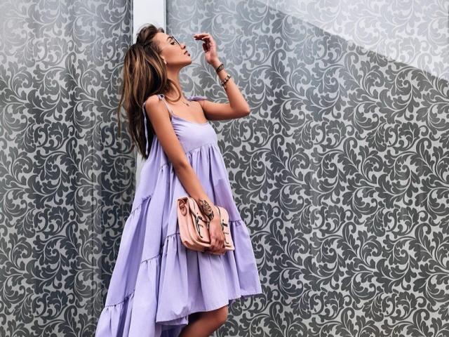 Kissamya with long brown hair, wearing a lilac dress, holding a pink clutch, touching her nose, tattoo on wrist, standing against a gray, ornate-patterned wall.