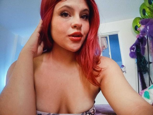 SophieHill with red hair, slightly parted lips, and a hand in her hair, showing her bare boobs in a selfie. She has a cute, sexy look.