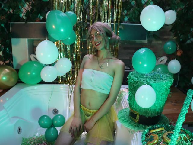 Allison Pearzon met roze haar, een witte top, een gele rok, zittend in een jacuzzi, groene en witte ballonnen, groene en gouden decoratie, lachend, handen op de dijen, kleine borsten..