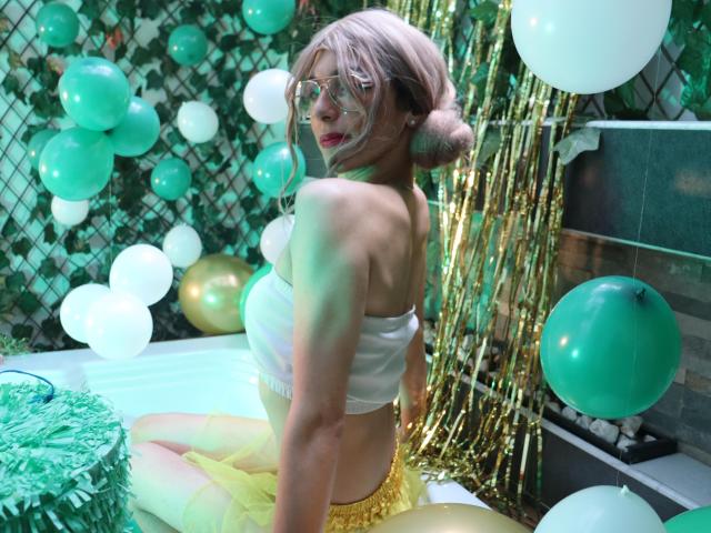 AllisonPearzon with pink hair in buns, wearing transparent strapless top, yellow skirt, and glasses, kneeling in green and white balloons, gold streamers, and green patterned wall.