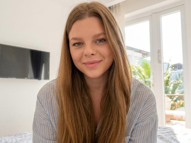RhonaBells has long, straight brown hair, blue eyes, and a slight smile. She's wearing a blue and white striped shirt, sitting in a bright room.